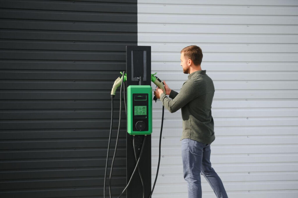 Mężczyzna dokonuje montażu stacji ładowania samochodów elektrycznych na tle ściany o dwóch kolorach.